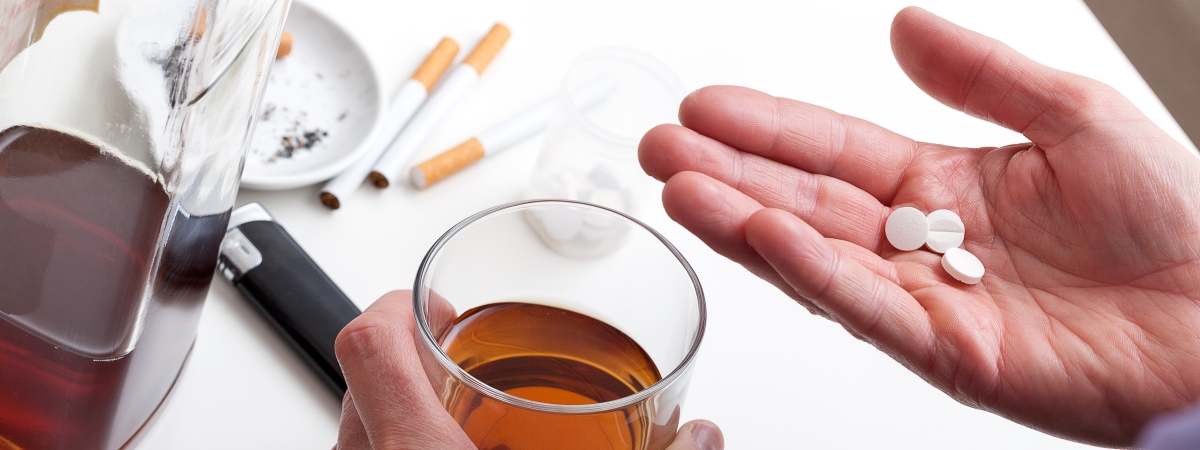 Eine Hand hält das mit Alkohol gefüllte Glas. In der anderen Hand liegen Medikamente. Auf dem Tisch liegen Zigaretten.