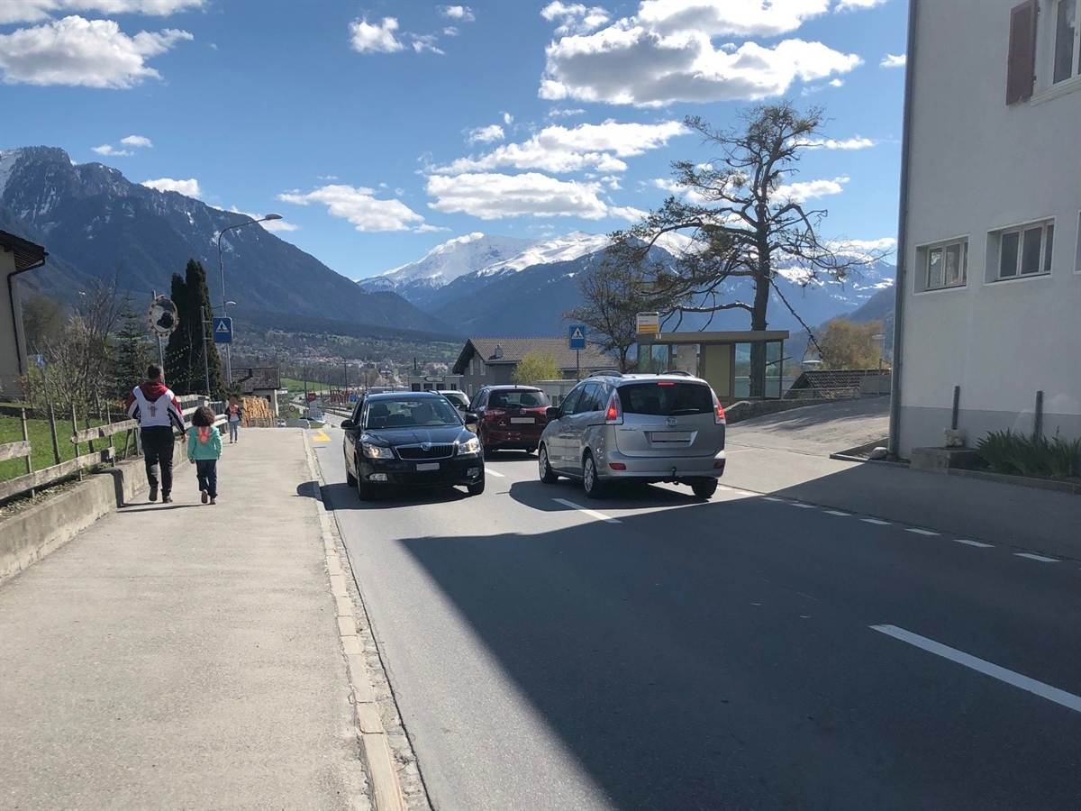 Drei in Richtung Trimmis und ein in Richtung Dorfzentrum Zizers fahrende Autos. Links davon ein Trottoir mit einem Erwachsenen und einem Kind sowie ein weiteres alleine beim Fussgängerstreifen stehendes Kind.