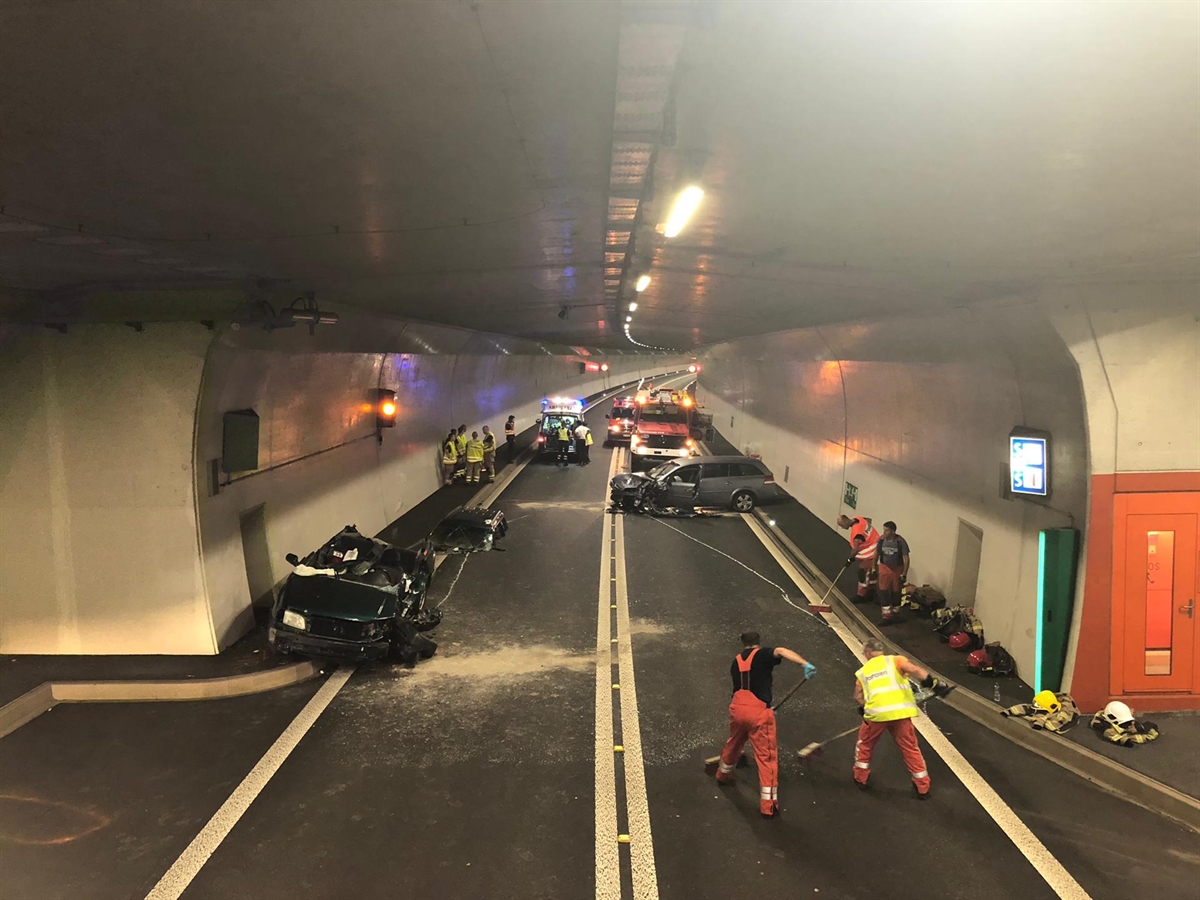 Aufnahme des Tunnels. Im Vordergrund Einsatzkräfte, die die Unfallstelle räumen. Links ein total beschädigtes Auto mit aufgeschnittenem Dach. Rechts ein querstehendes total beschädigtes Auto. Im Hintergrund Einsatzfahrzeuge.