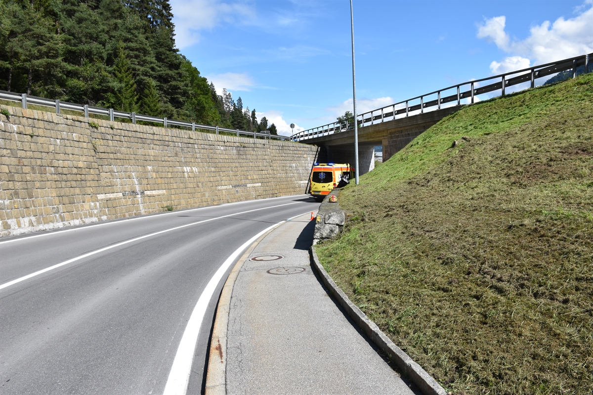 Rechts eine ansteigende Wiesenböschung. Links davon das Trottoir und die Strasse, welche eine Rechtskurve beschreiben. Im Hintergrund eine Ambulanz.