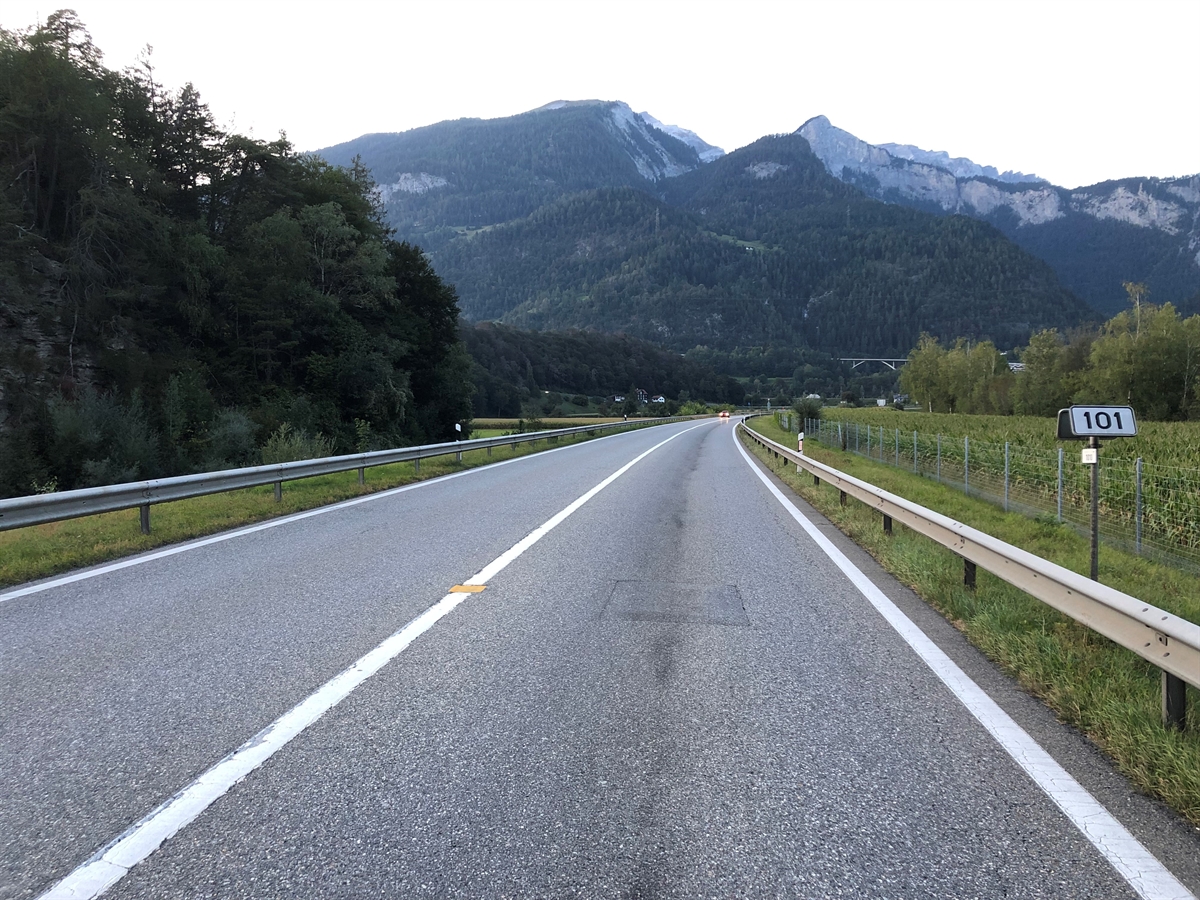 Die Strasse, links und rechts von Leitplanken begrenzt und anschliessend jeweils Wiesland. Im Hintergrund Berge.