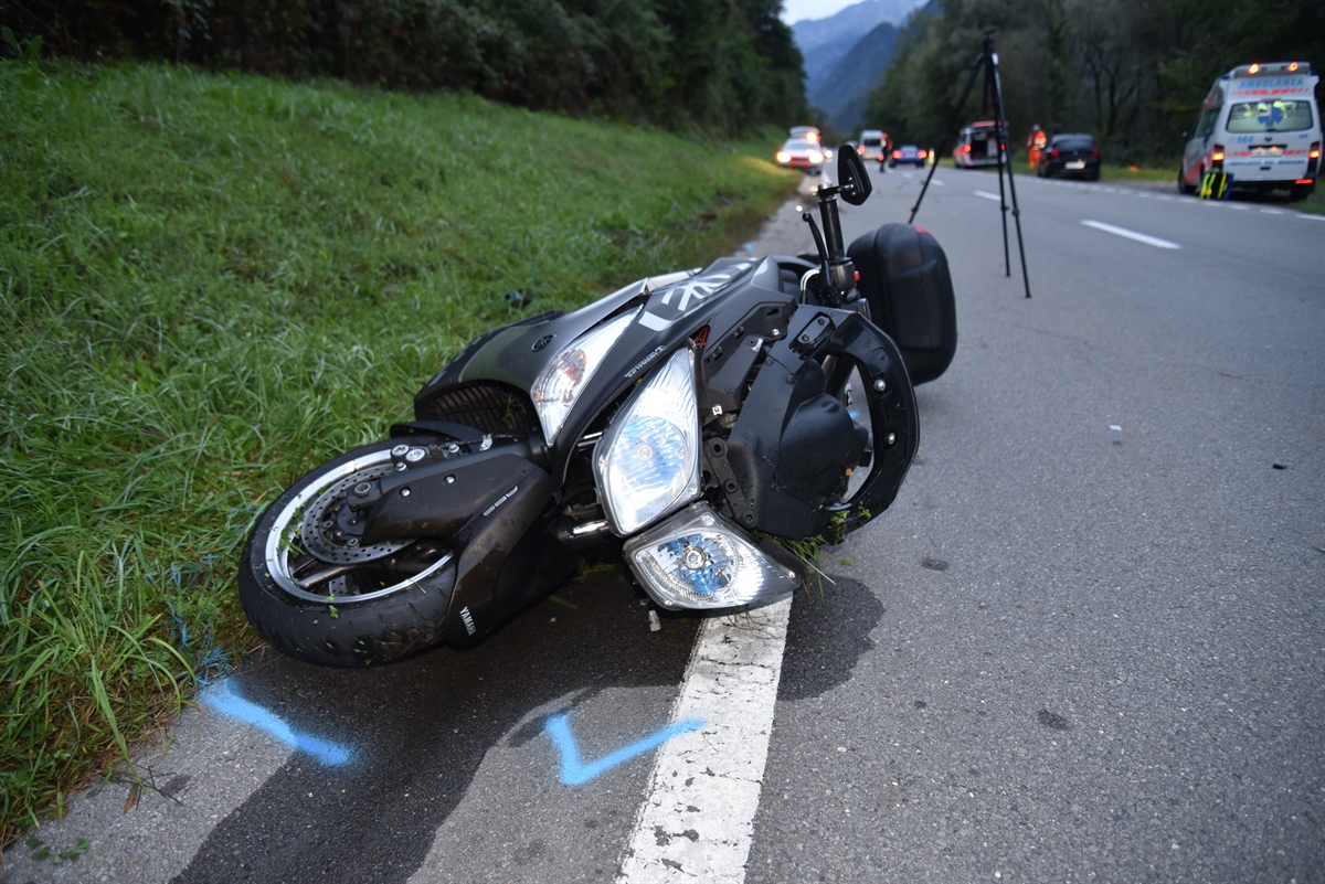 Nahaufnahme des am Strassenrand liegenden Rollers. Rechts die Strasse, an deren Fahrbahnrändern weitere Fahrzeuge – darunter Ambulanz und Polizeiauto – stehen.