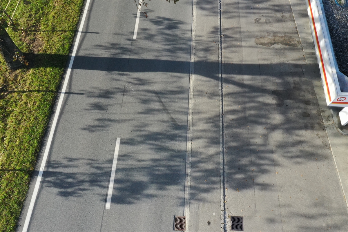 Luftaufnahme, links Wiese, mittig die Strasse mit Mittelmarkierung, rechts ein Vorplatz und das Dach der Tankstelle.