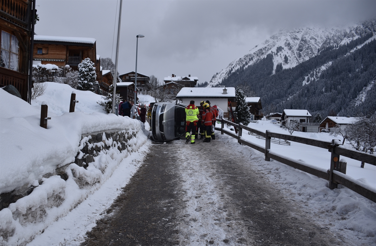 Unfallstelle bergwärts gesehen. Links ein Haus, rechts davon die Strasse. An deren Rand frontal das auf der Fahrerseite liegende Auto. Davor Rettungskräfte.