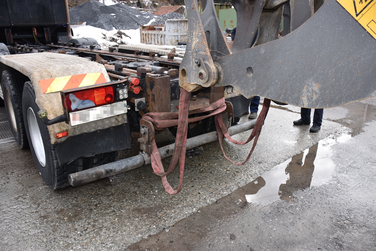 Rechts der Greifarm des Pneuladers, links das Lastwagenheck. Zwischen dem Pneulader und dem Lastwagen hängen Gurte.