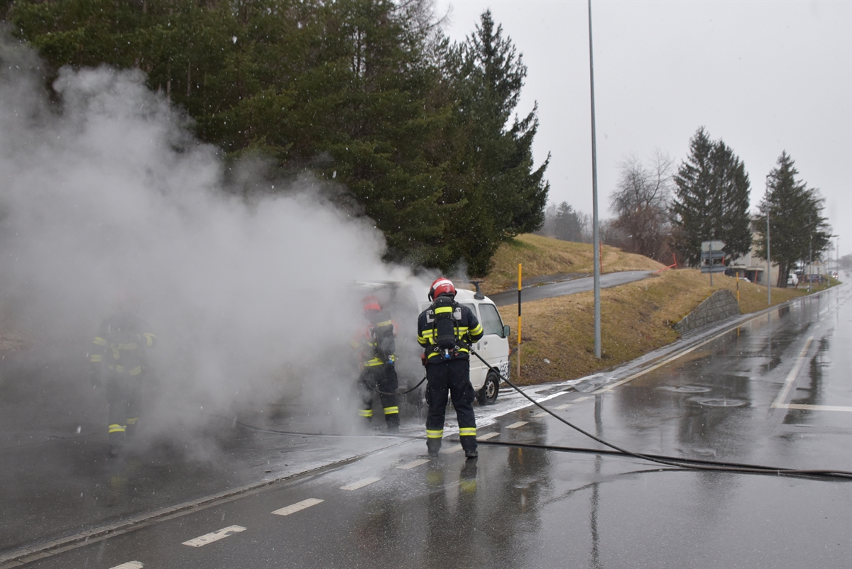 Links weisser Rauch, rechts davon auf einer Zufahrt der Lieferwagen und zwei mit Löscharbeiten beschäftigte Feuerwehrleute, rechts davon die Hauptstrasse.