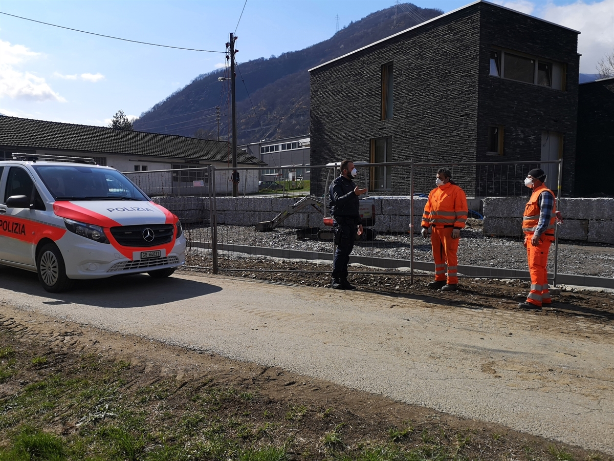 Links ein Polizeiauto, rechts davon ein Polizist, welcher sich mit zwei Bauarbeitern unterhält.