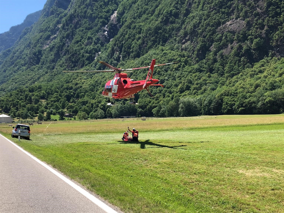 Links ein Stück Strasse, rechts davon eine Wiese mit auf einer Höhe von ca. 5 m schwebendem Rega-Helikopter und zwei Personen, welche darunter beim mit Bahre auf der Wiese abgelegten Verletzten kauern.