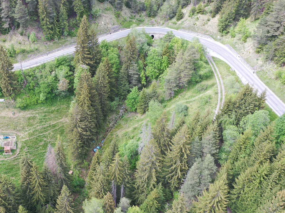 Drohnenaufnahme der Unfallörtlichkeit mit der langgezogenen Rechtskurve. Im Bachbett im Wald ist das hellblaue Unfallauto erkennbar