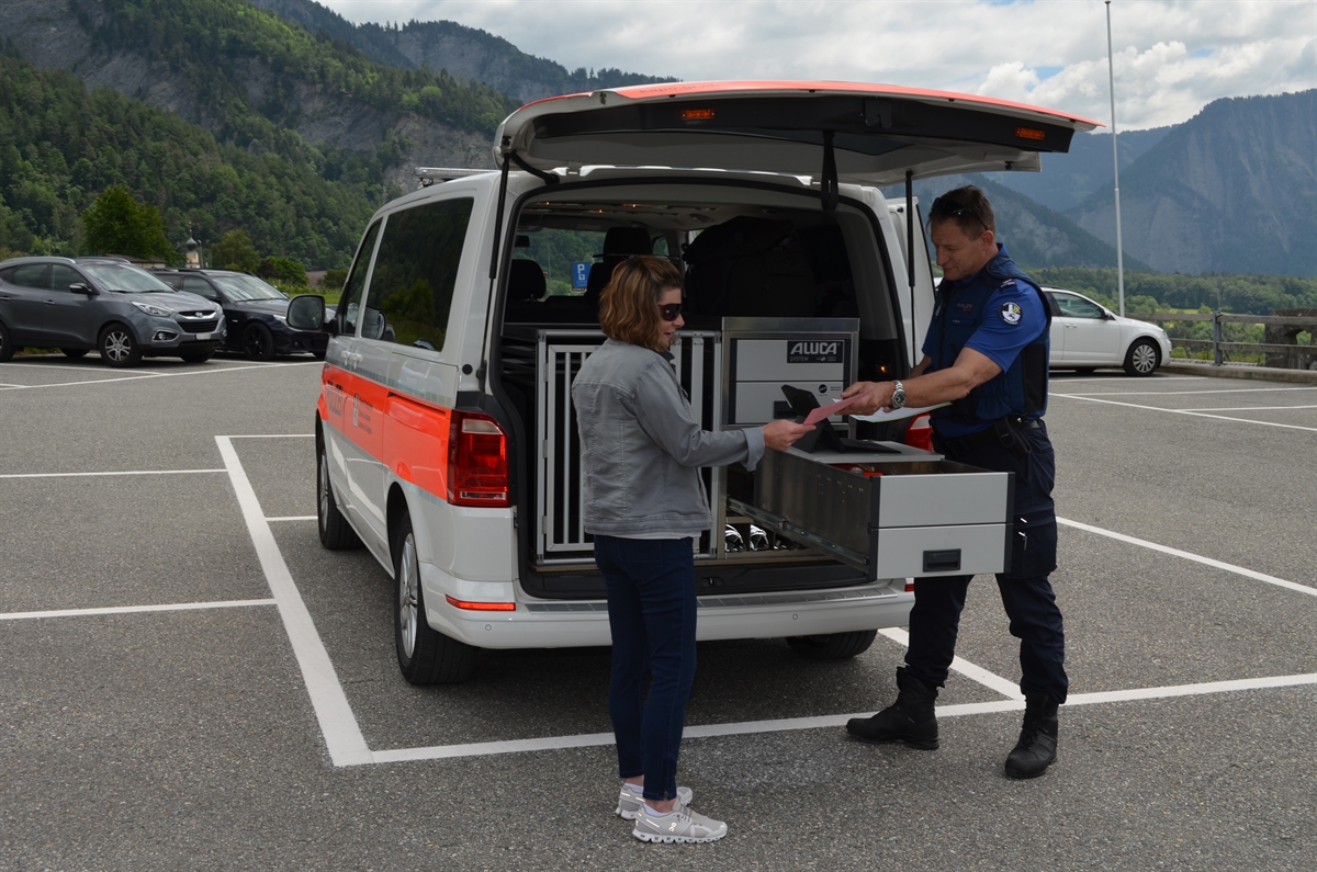 Ein Parkplatz mit einem Polizeiauto mit geöffneter Heckklappe. Am Heck steht eine Frau, welche von einem Polizisten Unterlagen überreicht erhält.