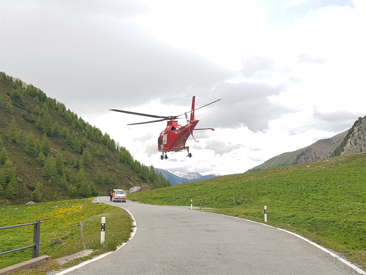 Die Umbrailstrasse mit der Rechtskurve. Über der Fahrbahn schwebt der Regahelikopter. Im Hintergrund stehen ein Polizeiauto und ein Polizist. Letztgenannter sperrt die Strasse.