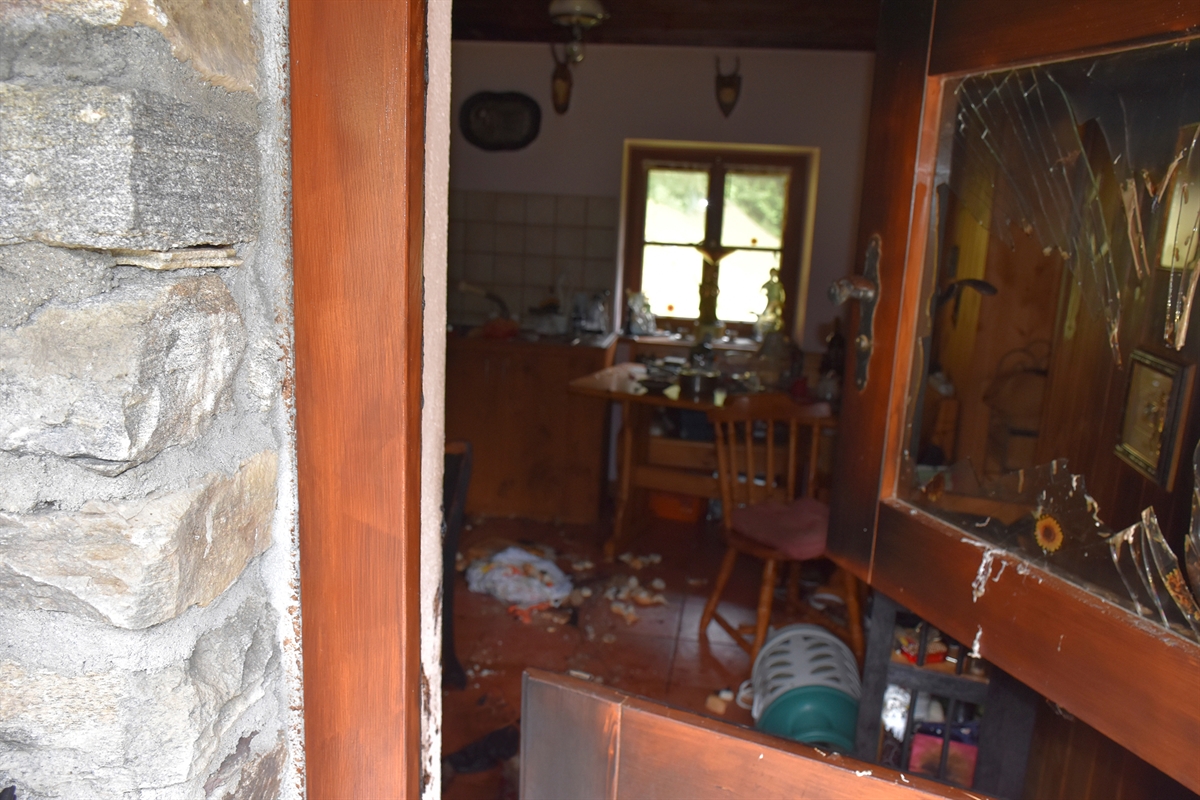 Blick von der Eingangstüre in das Maiensäss. Die horizontal zweigeteilte Türe ist im oberen Bereich weiter geöffnet. Das Fenster in diesem oberen Bereich ist zersplittert. Im Maiensäss liegt eine Gasflasche, es herrscht Unordnung. 
