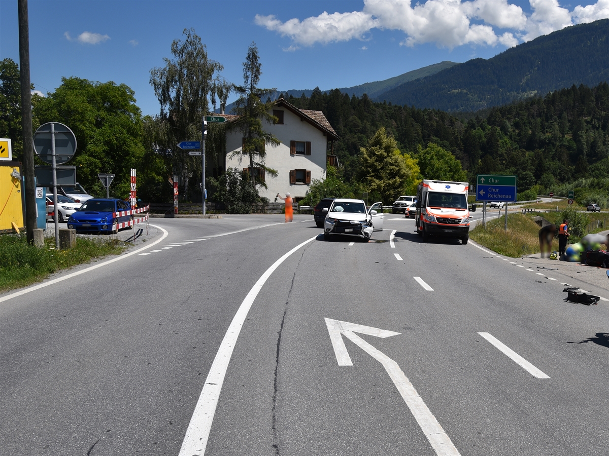Verzweigung in der Isla in Bonaduz. Ein Bauarbeiter regelt den Verkehr. Mitte im Bild das Unfallfahrzeug und rechts davon die Ambulanz. Am rechten Bildrand Personen die sich um den Verletzten kümmern sowie ein Polizist.