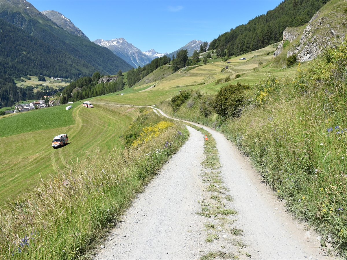 Feldweg in Richtung Ardez mit einer Ambulanz sowie zwei Polizeifahrzeugen auf der Wiese stehend.