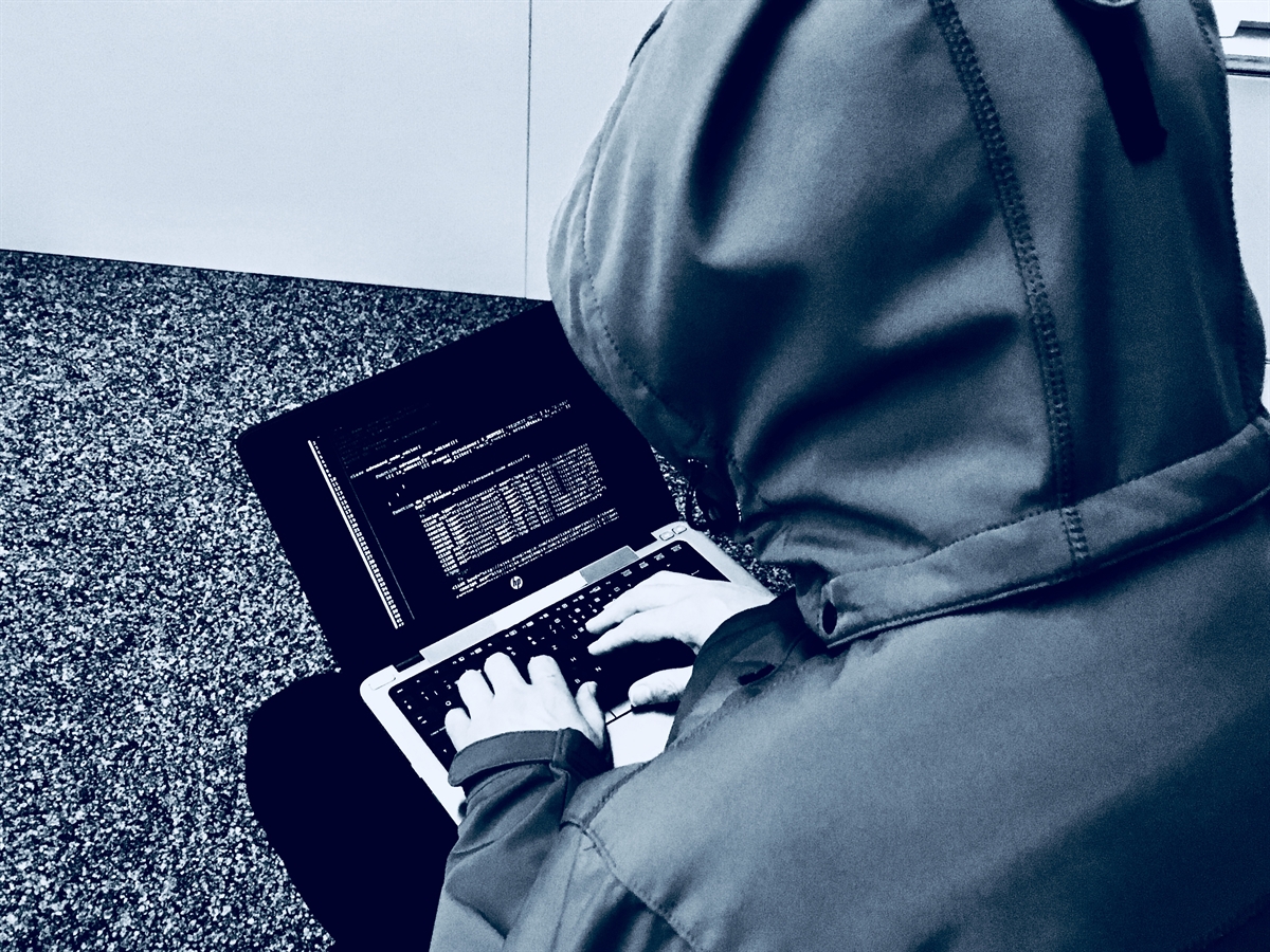Ein Hacker von hinten, Kapuze über den Kopf, auf den Beinen einen Laptop, die Finger auf der Tastatur.