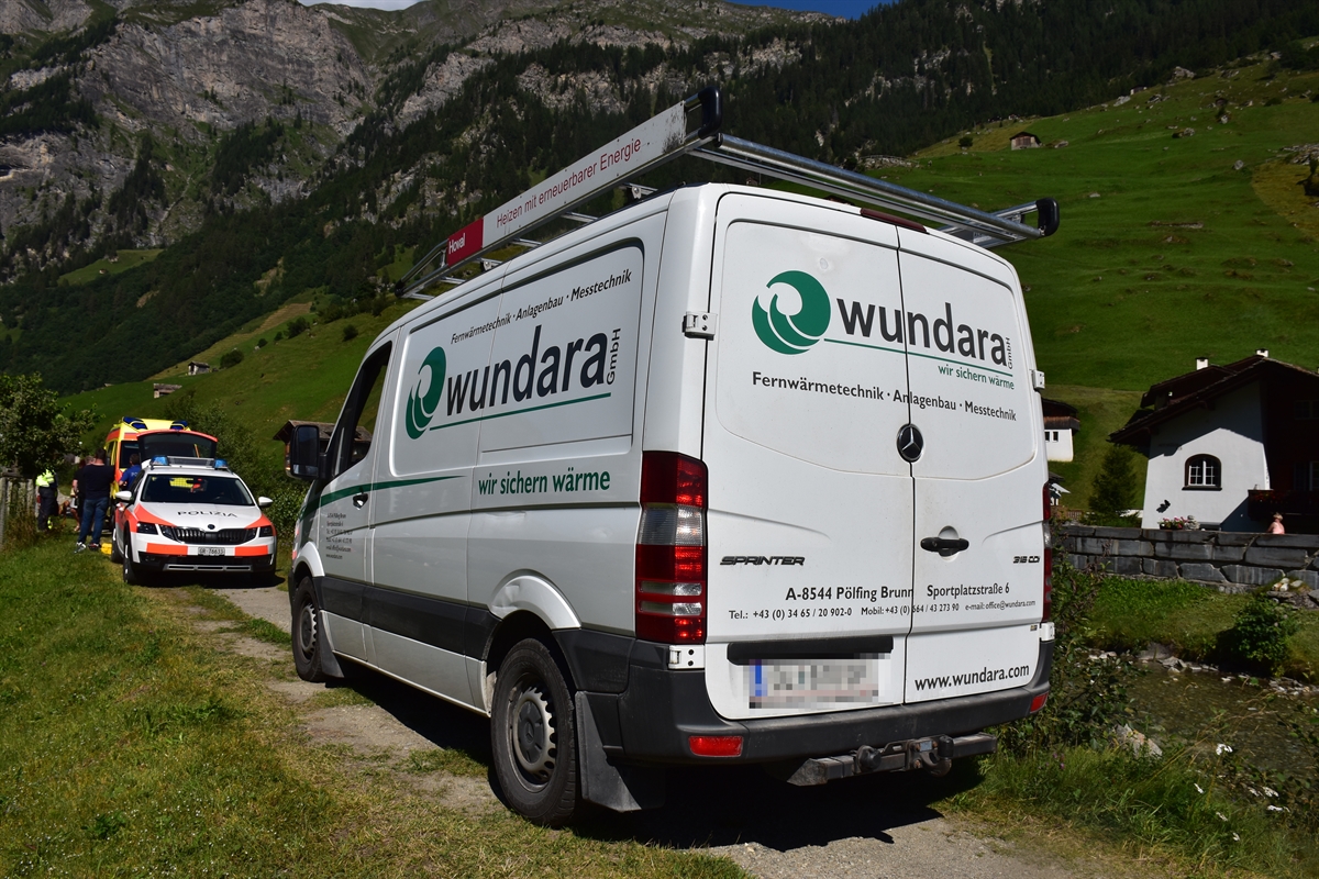 Weisser Lieferwagen seitlich von hinten auf Naturweg. Links und Rechts Wiesland. Im Hintergrund das Polizeifahrzeug und die Ambulanz 