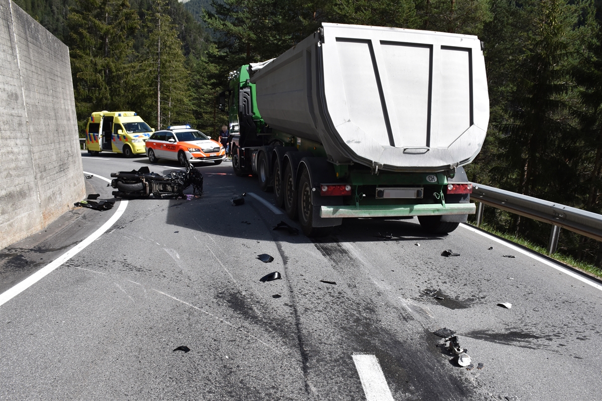 Links eine Stützmauer, rechts davon auf der Fahrbahn das total beschädigte Motorrad. Rechts davon das Sattelmotorfahrzeug von hinten. Im Vordergrund Kollisions- und Kratzspuren auf der Fahrbahn, im Hintergrund ein Polizeiauto und ein Krankenwagen.