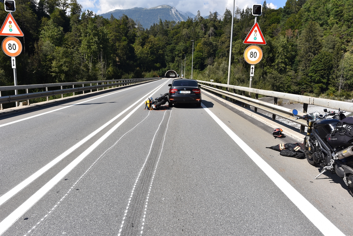 Geradlinige nicht richtungsgetrennte Autostrasse in Richtung Plazzas Tunnel gesehen. Auf der rechten Fahrspur steht ein Personenwagen und am hinteren linken Fahrzeugecken liegt das verunfallte Motorrad am Boden. Am Strassenrand rechts steht ein zweites nichtbeteiligtes Motorrad.