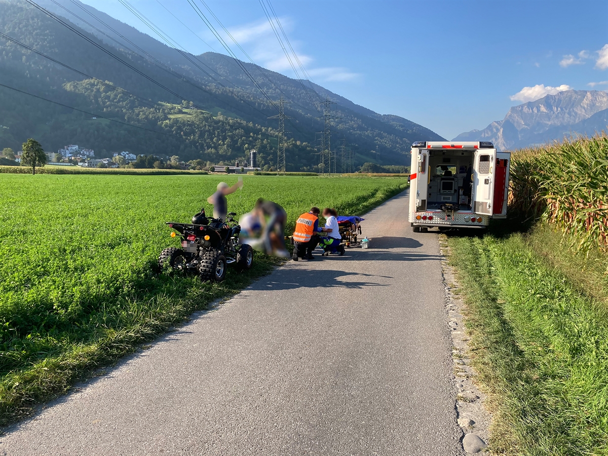 Feldweg in der grünen Landschaft. Links vom Weg das Vierradfahrzeug und die Rettungskräfte mit dem Verletzten. Rechts am Wegrand grenzt ein Maisfeld. Die Ambulanz der Rettung Chur ist im Hintergrund sichtbar.