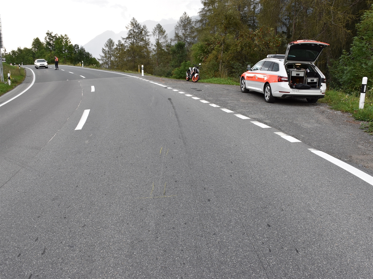 Linkskurve mit Brems- und Kratzspuren auf der Fahrbahn. Rechtsseitig steht das Motorrad sowie ein Patrouillenfahrzeug der Kantonspolizei Graubünden. Am linken Bilderrand wird der Verkehr durch einen Polizisten angehalten