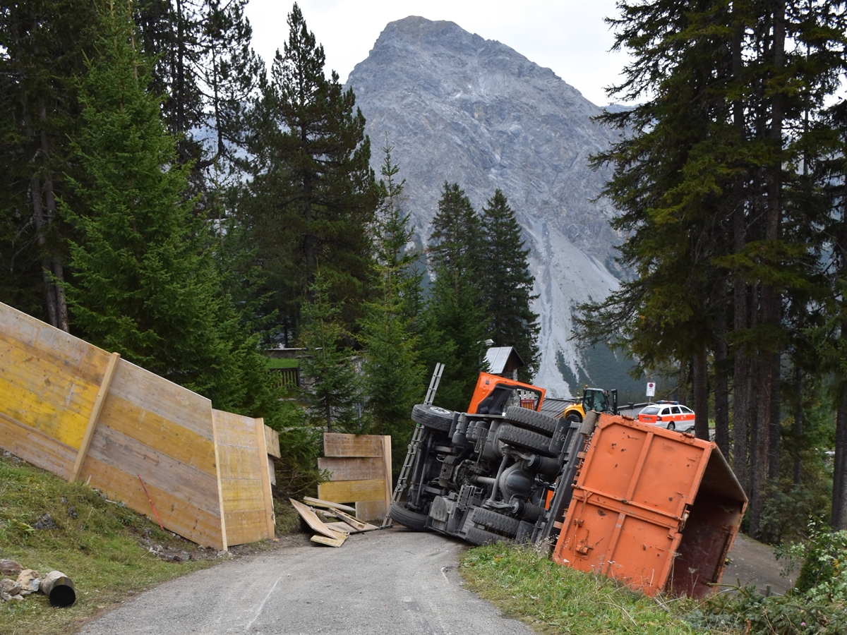 Die abfallende Strasse. Auf dieser sowie auf dem rechts angrenzenden Wiesenbord liegt ein Lastwagen auf der rechten Seite. Das zentrale Geschehen ist umsäumt von Bäumen, einem Polizeiauto und einem Berg.