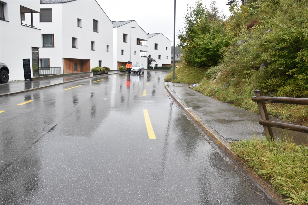 Die Unfallstelle bergwärts gesehen, im Hintergrund ein Polizist. Links der Strasse Häuser und rechts der zur Strasse führende Fussweg.