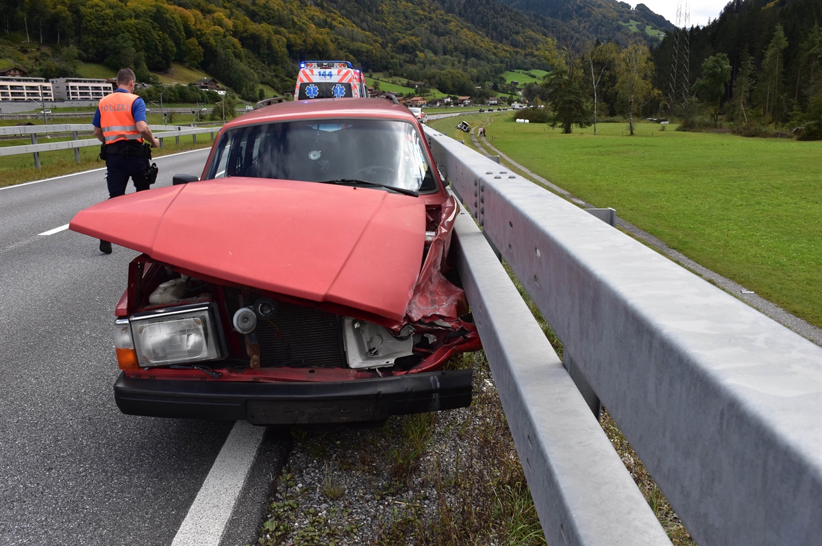 Frontaufnahme des talauswärts gefahrenen total beschädigten Autos an der Leitplanke. Im Hintergrund ein Polizist und eine Ambulanz.