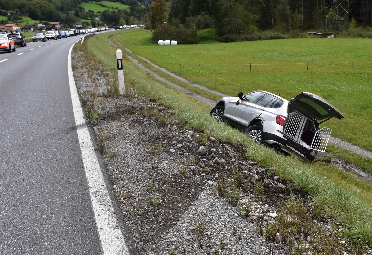 Links die Strasse und rechts die abfallende Wiesenböschung in welcher das zweite Auto steht. Auto von hinten schräg links, die linke Seite und hintere Radaufhängung stark beschädigt.