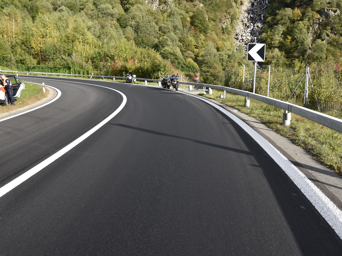 Manzei-Kehre mit Ölspur auf der Bergspur der A13. Zwei Motorräder stehen auf den Seitenständern und das verunfallte Motorrad lehnt an der Leitplanke. Am linken Bildrand öffnet ein Kantonspolizist die hintere Türe eines Patrouillenfahrzeuges.