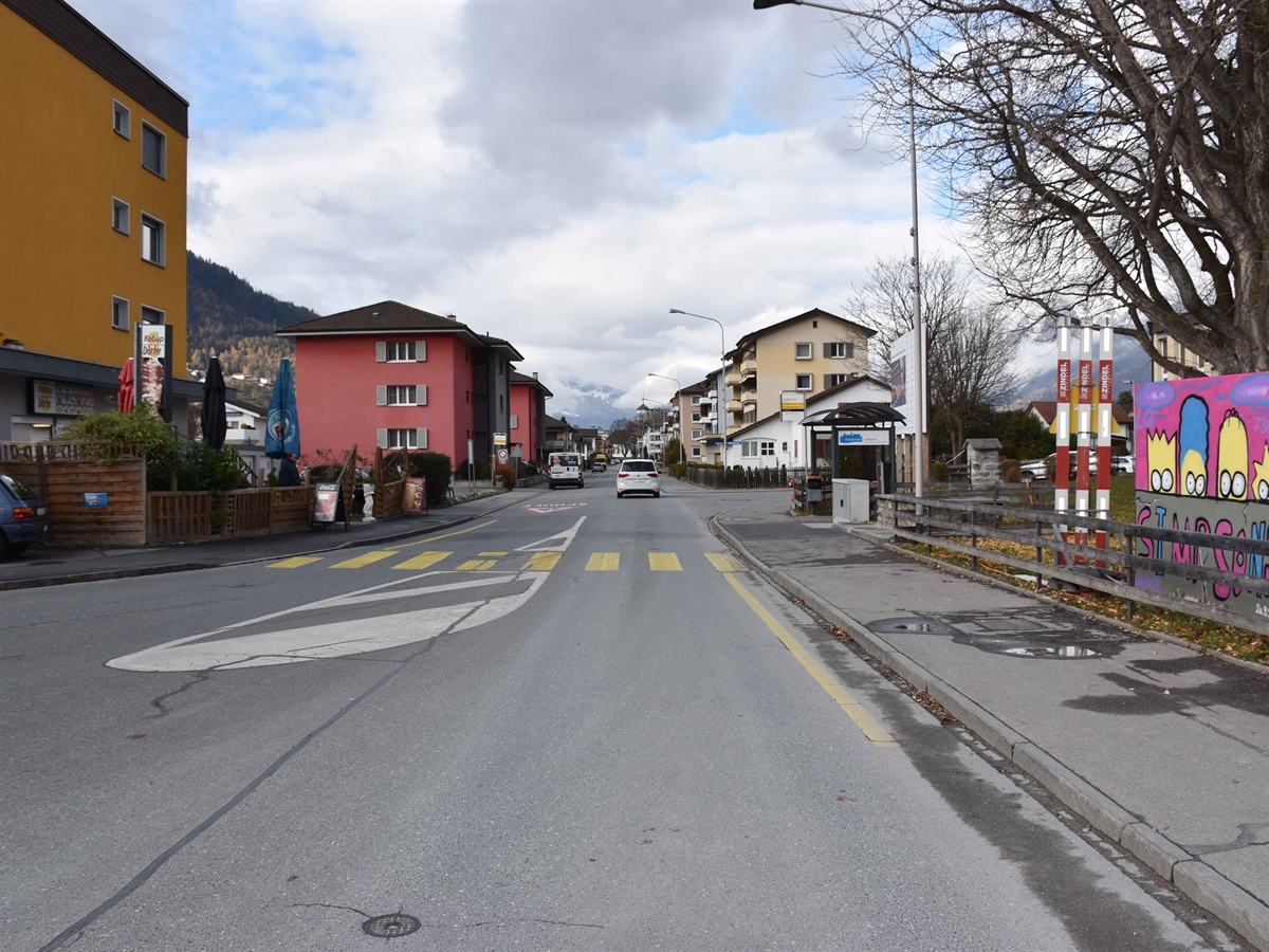 Aufnahme in Richtung Dorfzentrum. Rechts und links jeweils ein Trottoir. Inmitten einer Sperrfläche ein Fussgängerstreifen. Die Strasse ist von Häusern umsäumt.
