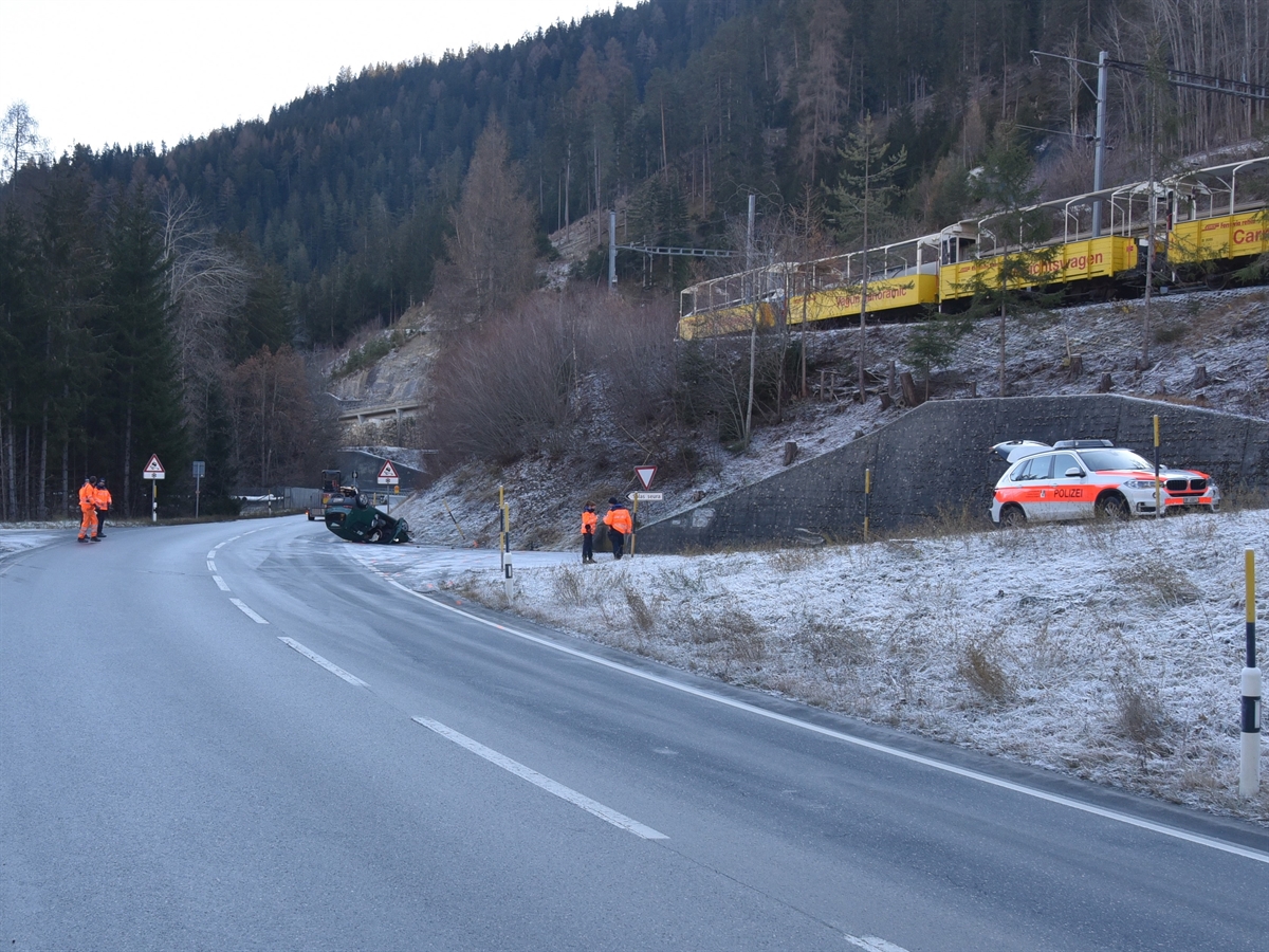 Unfallkurve in Fahrtrichtung gesehen. Rechts in Zufahrtsstrasse 1 Polizeiauto, 1 Polizistin und 1 Polizist. Oberhalb gelbe Aussichtswagen der RhB. Links aus der Ferne das auf dem Dach liegende Auto.