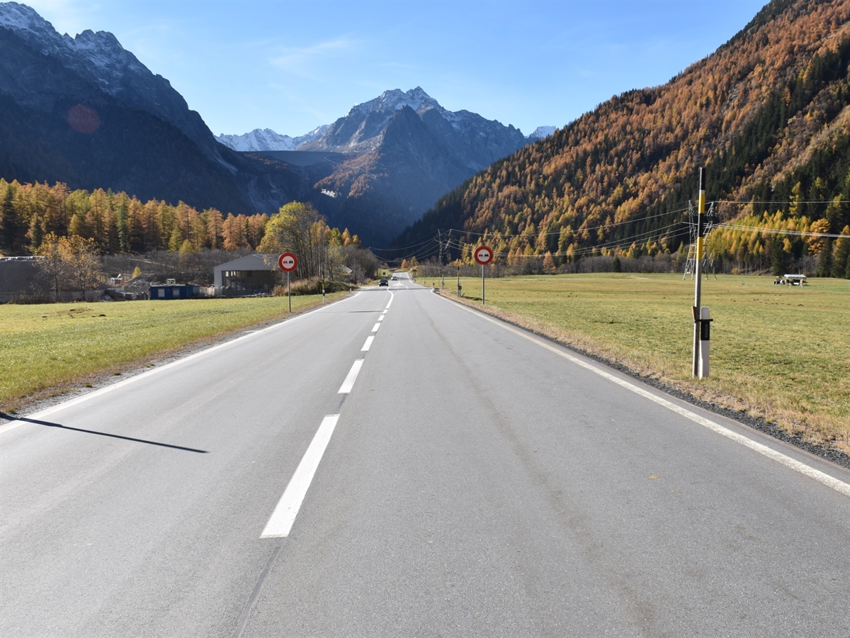 Aufnahme der Strasse, links und rechts umsäumt von Wiesen. Frontal im Hintergrund Blick auf Berge.