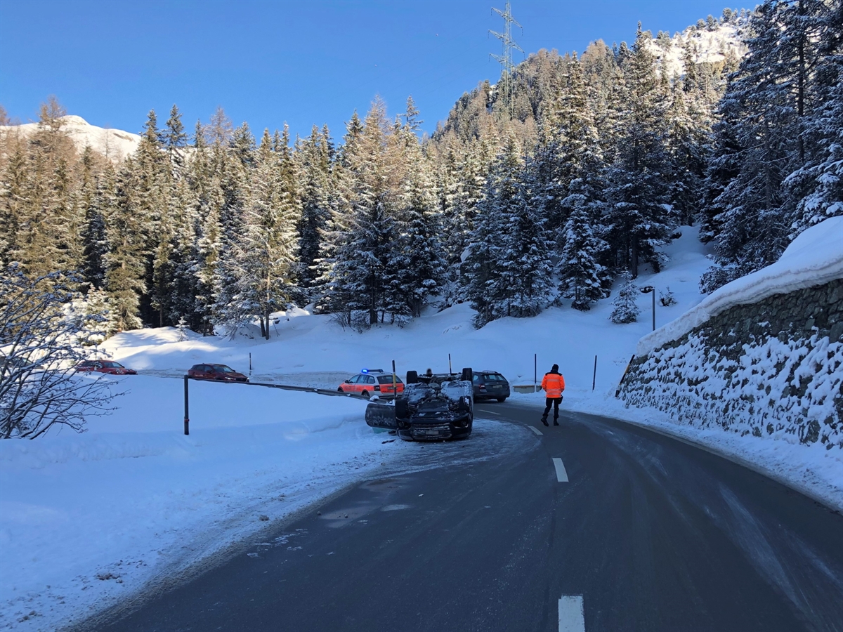 Unfallort aus der Gegenrichtung mit dem Unfallfahrzeug auf der linken Fahrbahn auf dem Dach liegend. Ein Polizist steht in der Mitte der Fahrbahn. Winterlandschaft mit blauem Bündnerhimmel.
