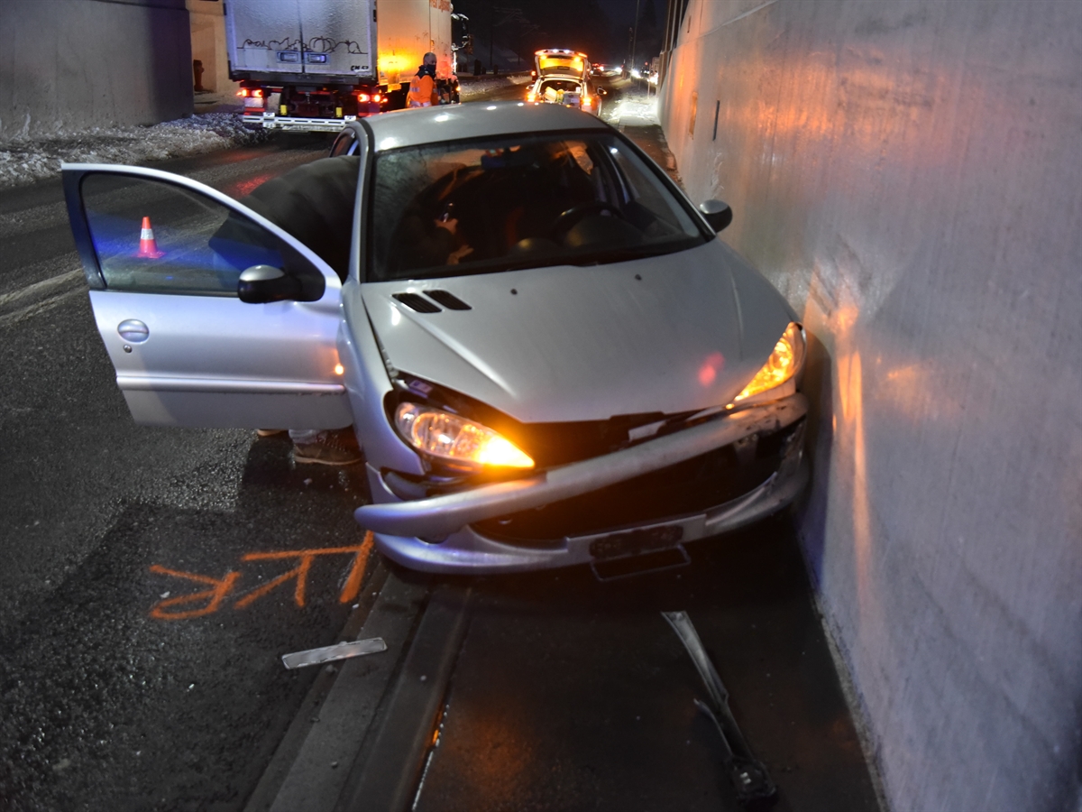 Rechts die Tunnelwand. Links davon frontal gesehen das beschädigte Auto. Im Hintergrund sieht man aus dem Tunnel auf die winterliche Strasse. Innerhalb des Tunnels ein Polizist und ein Polizeiauto.