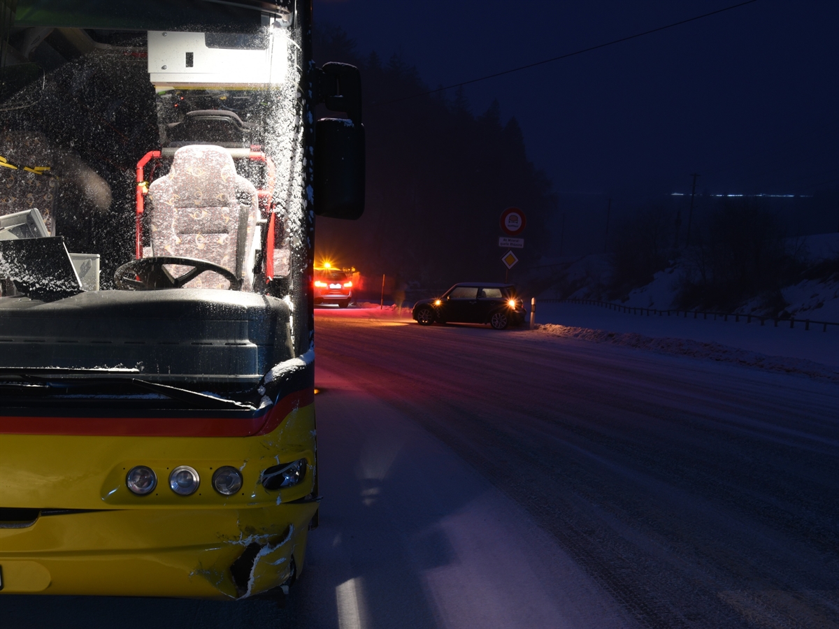 Aufnahme in der Morgendämmerung. Rechts im Hintergrund das Auto, links davon ein Polizeiauto mit Gelblicht. Im Vordergrund links frontal gesehen das Postauto mit beschädigter Front. 