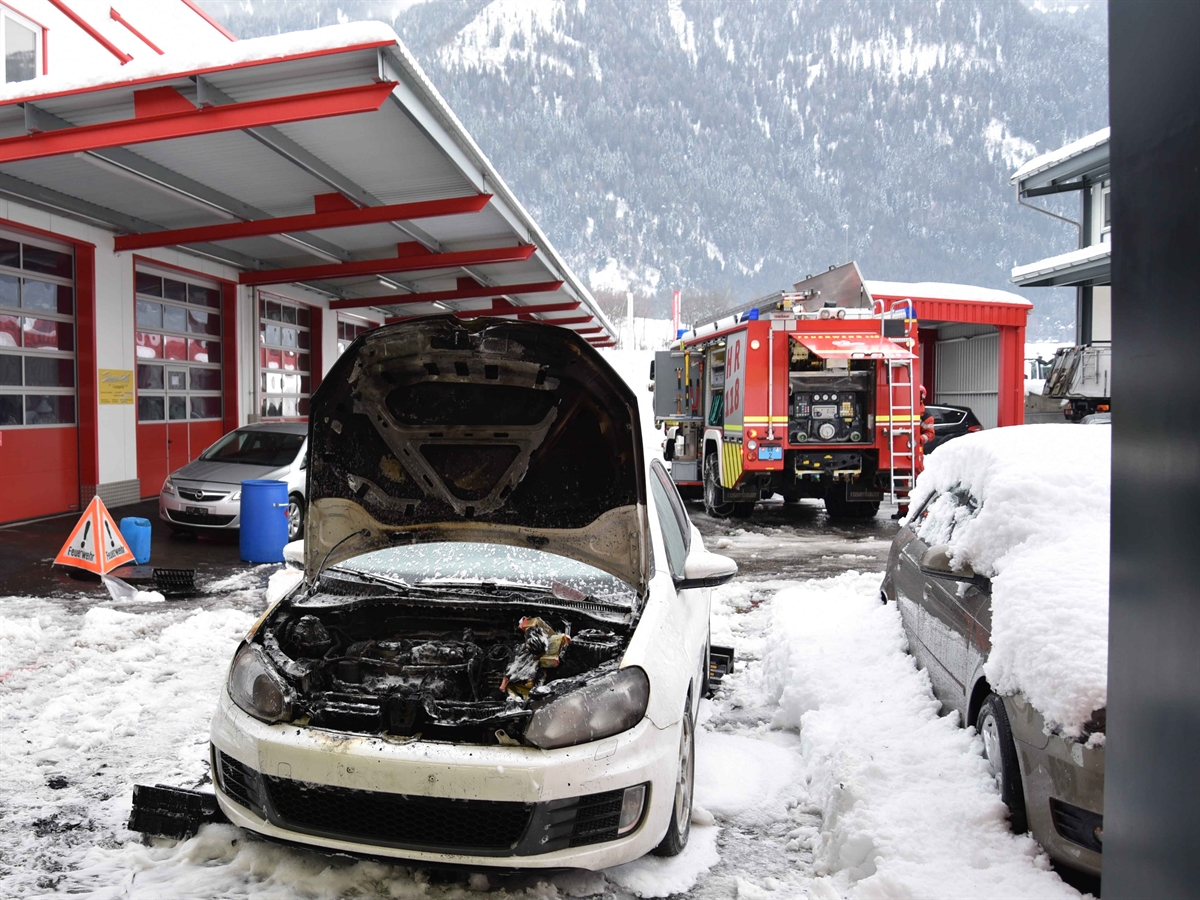 Blick aus der Halle auf das Auto. Die Haube ist offen, Brandschaden im Motorraum. Links dahinter im Hintergrund ein weiterer Gewerbebetrieb. Rechts im Hintergrund ein Feuerwehrauto.