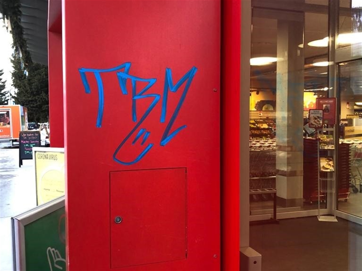 Ein roter Gebäudepfeiler, welcher mit einem blauen Schriftzug besprayt ist. Rechts im Hintergrund der Eingangsbereich zu einem Detailhandelsgeschäft.