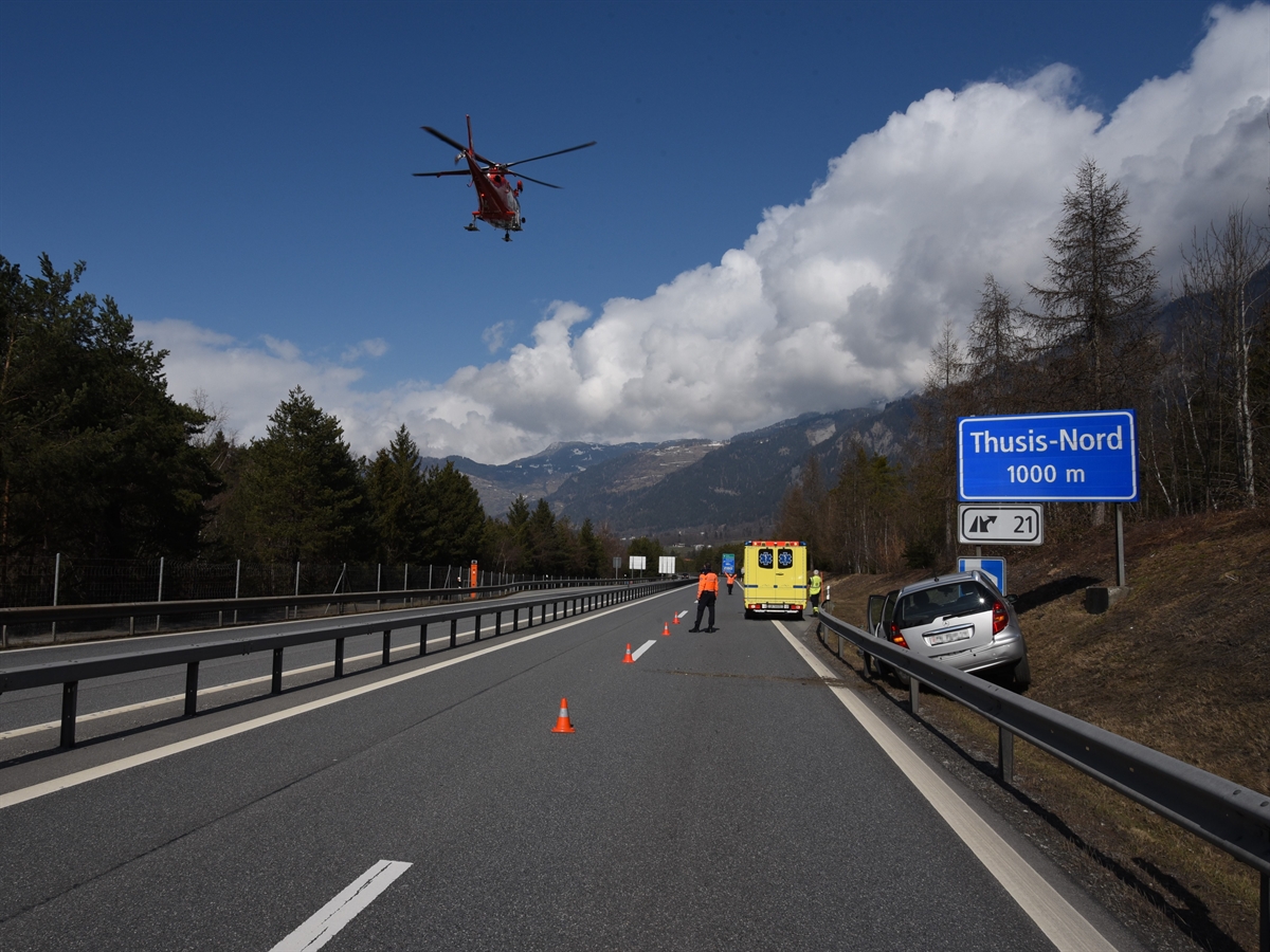 Die A13 in Richtung Norden. Rechts neben den Fahrspuren das Auto in Endlage. Auf der Strasse ein Rettungswagen, ein Polizist und eine Polizistin, welche den Helikopter – oben im Bild vor blauem Himmel – schulbuchmässig einweist.