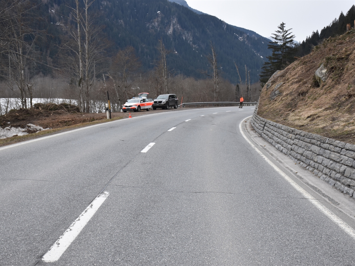 Der Verlauf der Oberalpstrasse vor einer Rechtskurve. Links auf einem Ausstellplatz stehen ein Patrouillenfahrzeug sowie das Fahrzeug des Wildhüters. Auf der Fahrbahn steht ein Polizist und hält den Verkehr an.