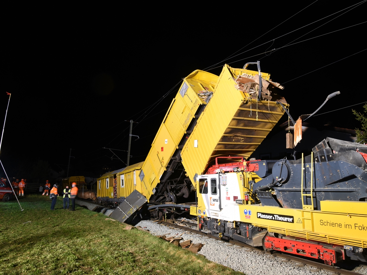Zwei aufgeschobene Bahnwagen liegen über einem Schottereinbauwagen. Links im Bild stehen Einsatzkräfte der RhB, Polizei sowie der Feuerwehr