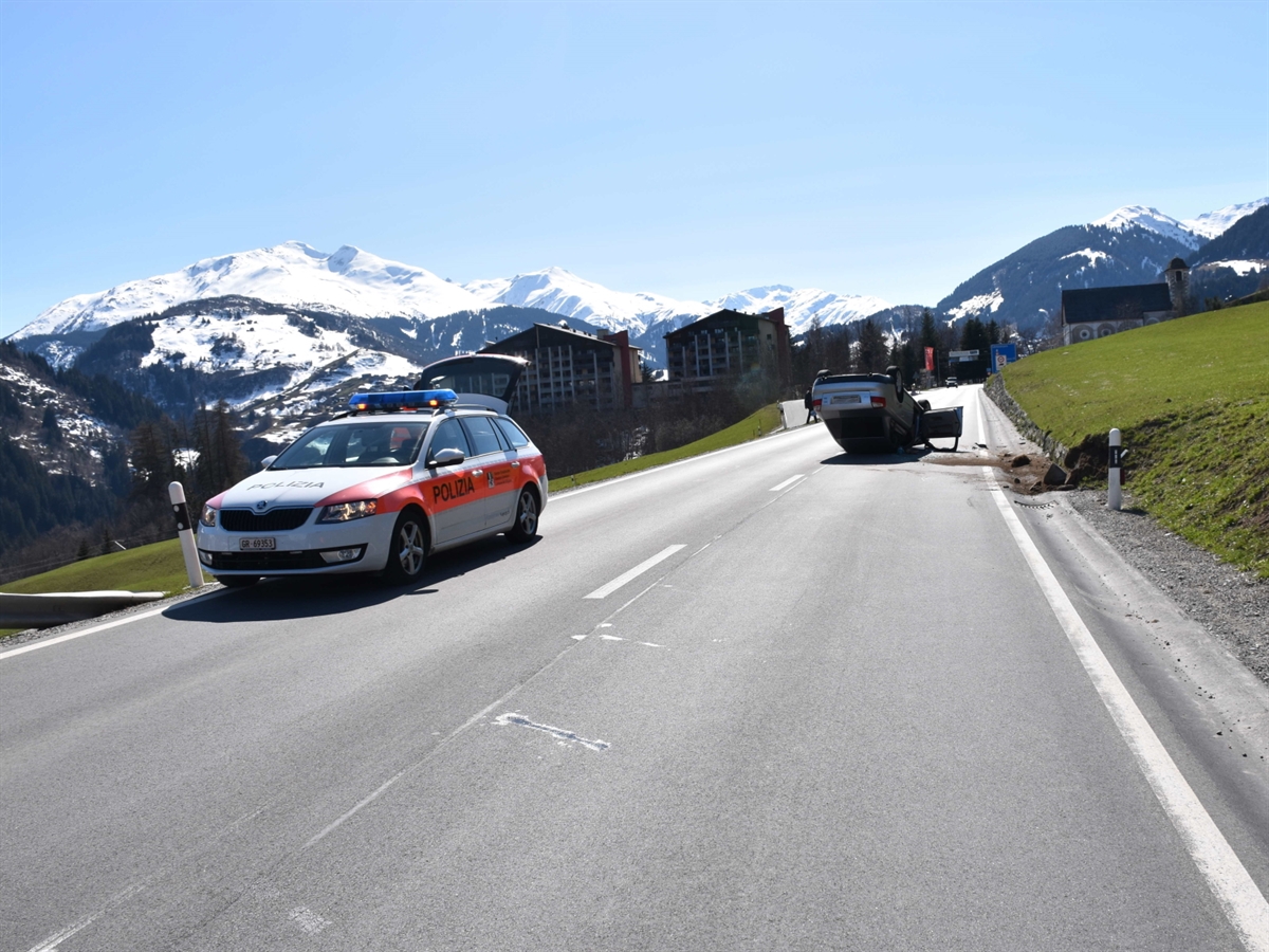 Die Gerade in Richtung Disentis/Mustér. Im Hintergrund Berge und blauer Himmel. Im Vordergrund links ein Polizeiauto und rechts ein wenig im Hintergrund das auf dem Dach liegende Auto.
