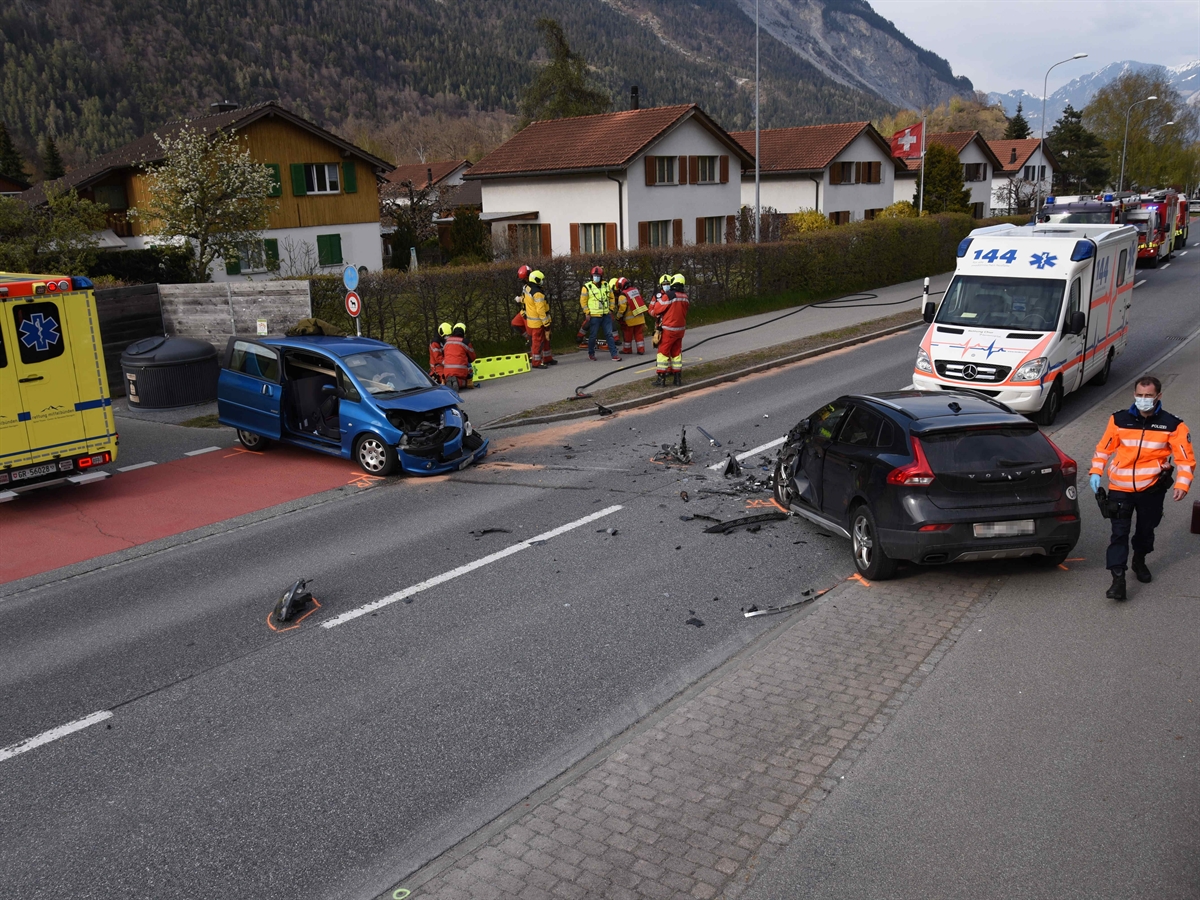 Die Unfallstelle mit von links gesehen: Eine Ambulanz, ein beschädigtes Auto, Feuerwehrleute, ein weiteres beschädigtes Auto, eine Ambulanz, im Hintergrund Feuerwehrautos und ganz rechts ein Polizist.