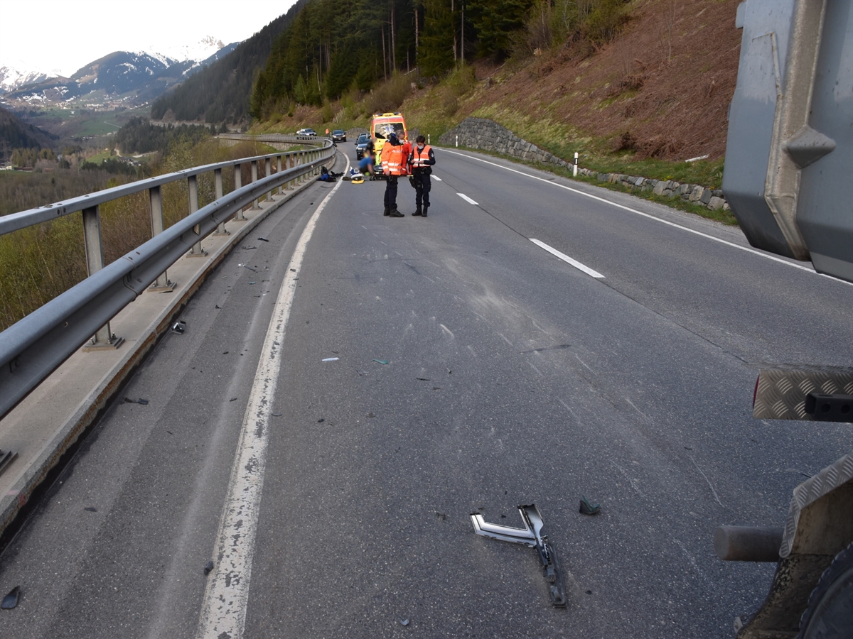 Am rechten Bilderrand sieht man noch das Heck des Lastwagens. Auf der Fahrbahn sind verschiedene Brems- und Schleifspuren. Im Hintergrund ein Ambulanzfahrzeug sowie Personen die eine Person auf einer Bahre betreuen. Vor diesen Personen zwei Polizisten die sich über das Spurenbild unterhalten.