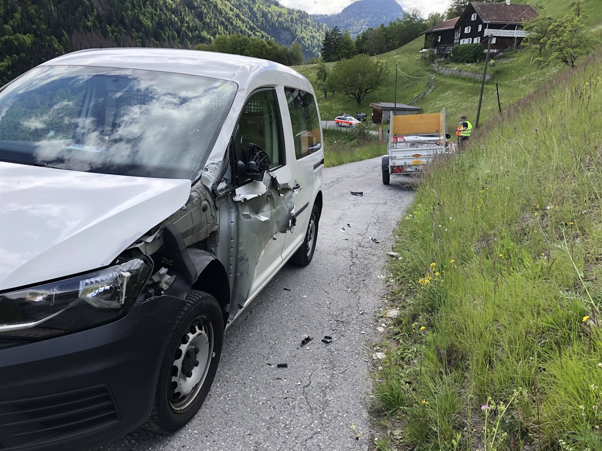 Die schmale Strasse, im Hintergrund der Anhänger von hinten. Im Vordergrund frontal das beschädigte bergwärts gefahrene Auto. Rechts davon die die ansteigende Wiesenböschung. 