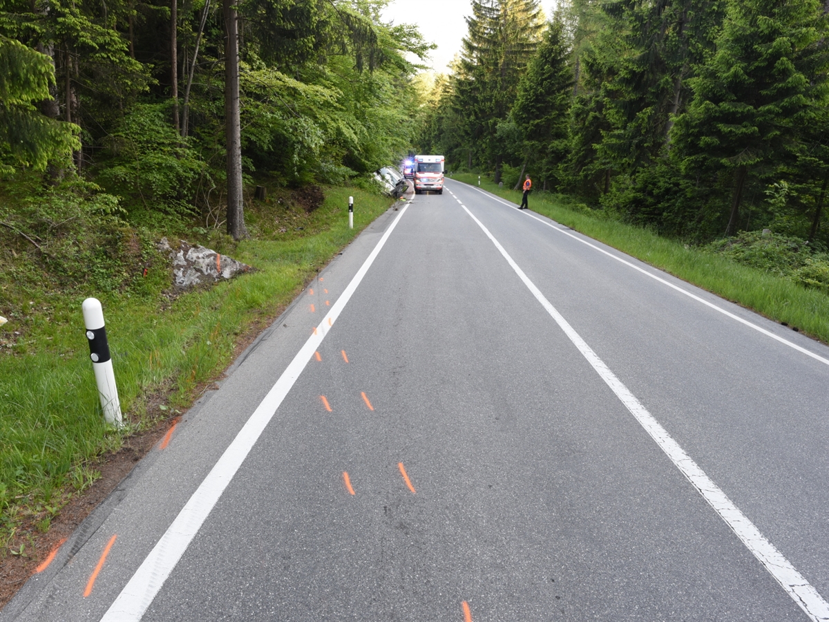 Blick aus der Unfallkurve in Fahrtrichtung gesehen. Die Strasse wird links und rechts von Wald begrenzt. Spuren, welche links von der Fahrbahn in die Böschung weisen und weiter unten das total beschädigte Auto an der Böschung. Dahinter Einsatzfahrzeuge.