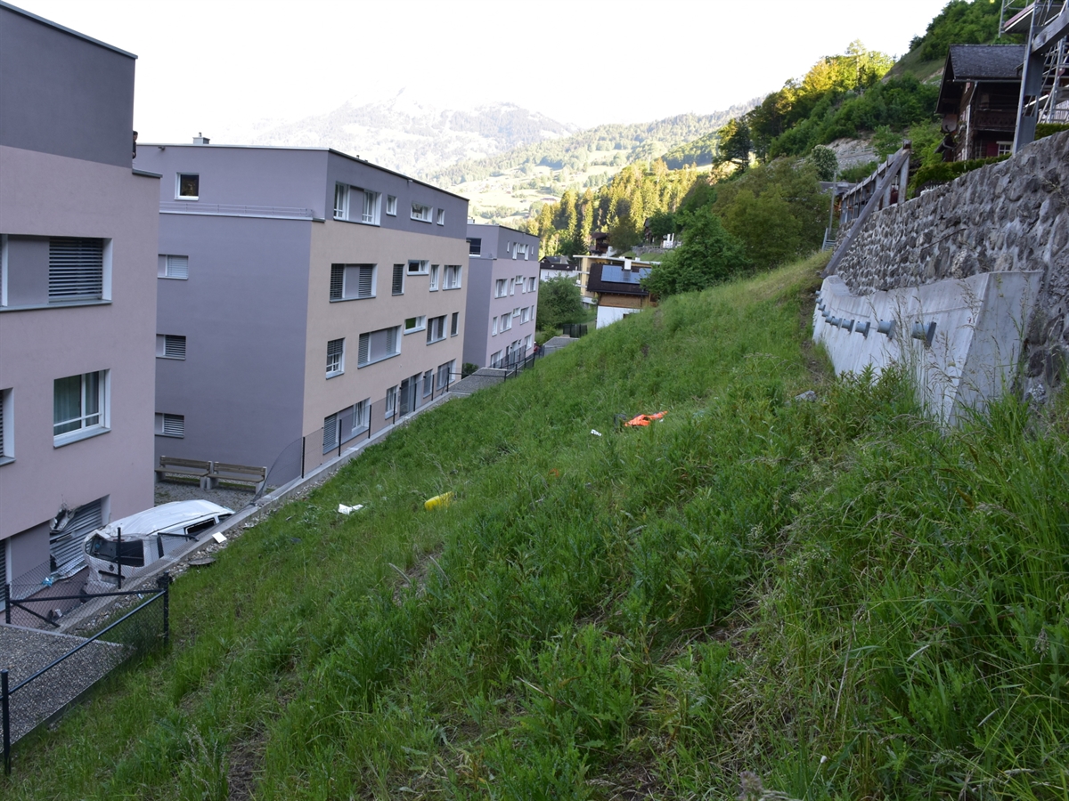 Wiese mit rechtsseitiger Strasse oberhalb einer Stützmauer. Am linken Ende der abfallenden Wiese liegt der Lieferwagen eingeklemmt zwischen Haus sowie einer weiteren Stützmauer. Gut sichtbarer Schaden an der Hausfassade der Parterrewohnung.