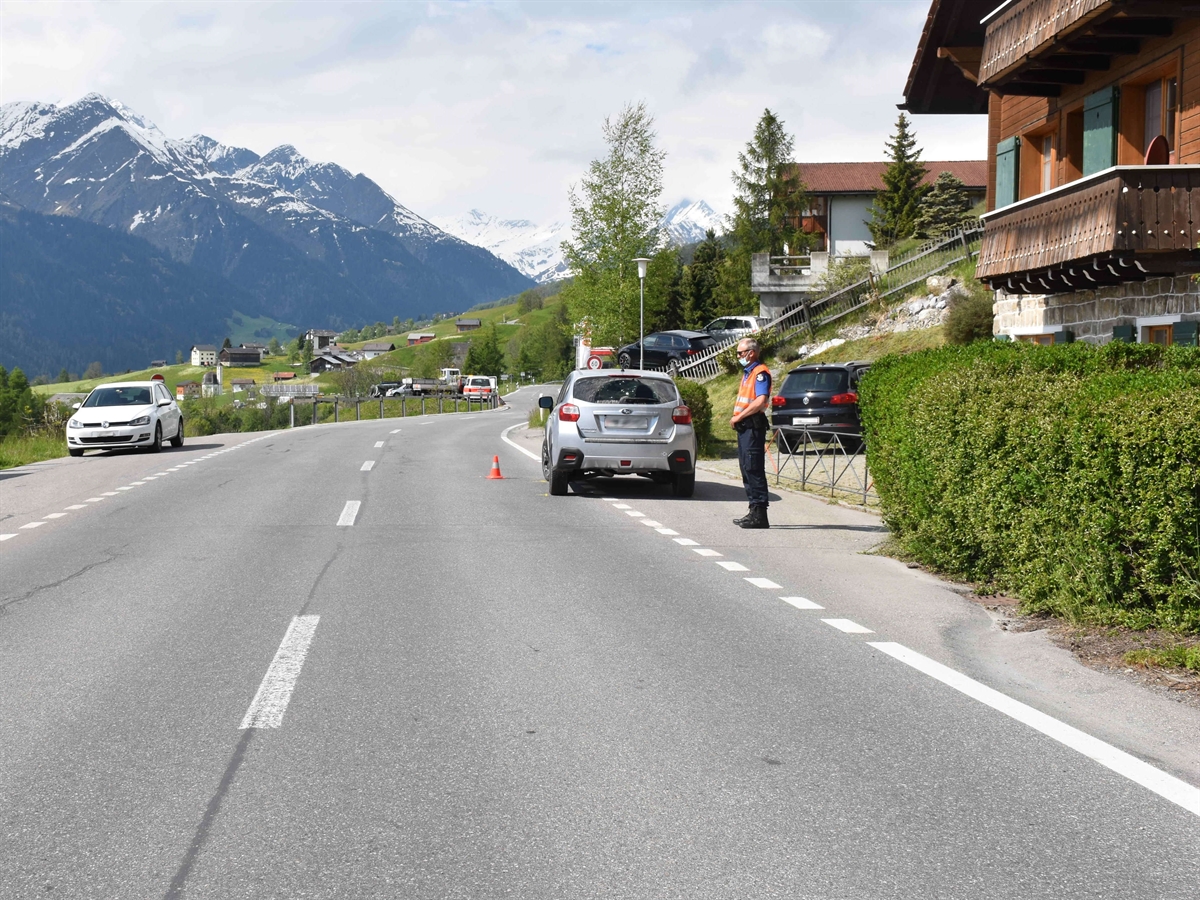 Rechts ein Haus mit einem Vorplatz. Links davon die Lugnezerstrasse an deren rechtem Rand das Auto steht. Weiter auf dem Bild ein Polizist, links gegenüber ein weiteres Auto sowie im Hintergrund Berge.