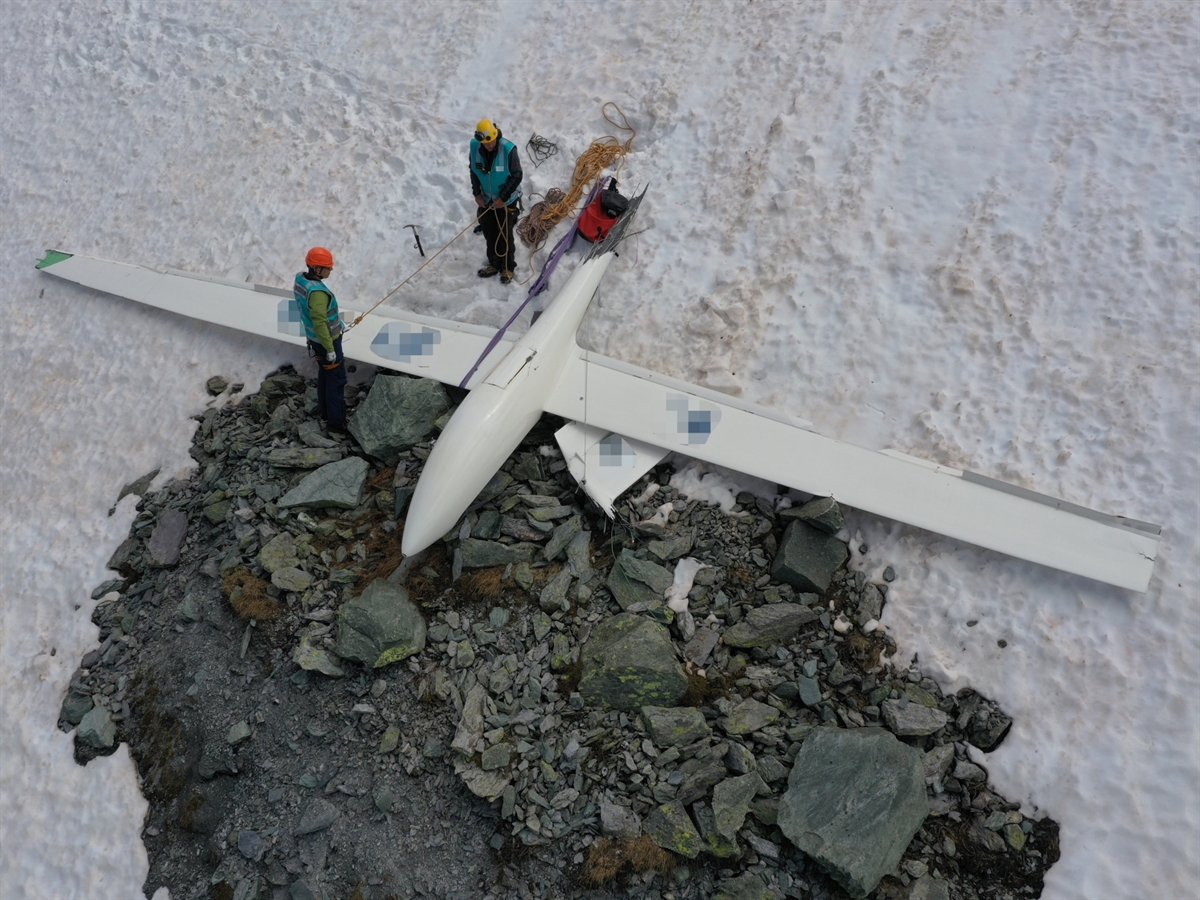 Das am Ende eines Schneefeldes liegende Segelflugzeug mit dem Rumpf nach oben. Zwei Einsatzkräfte beim Wrack.