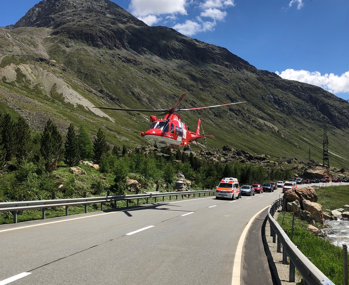 Rega Helikopter der gerade startet in der Mitte der Strasse. Dahinter sind zwei Polizeifahrzeuge und die Kolonne der gestauten Fahrzeuge. Blick Richtung Silvaplana. Rechtsseitig ist ein Bach.