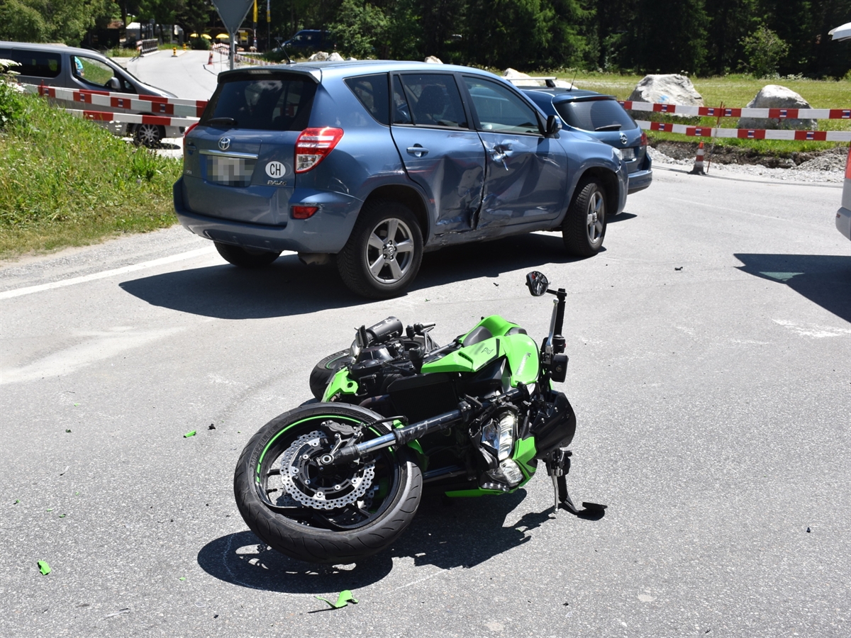 Die Verzweigung bei der Olympiaschanze in St. Moritz. Im Vordergrund liegt das beschädigte grüne Motorrad auf der linken Seite. Im Hintergrund steht das blaue Auto mit stark eingedrückter rechter Fahrzeugseite.